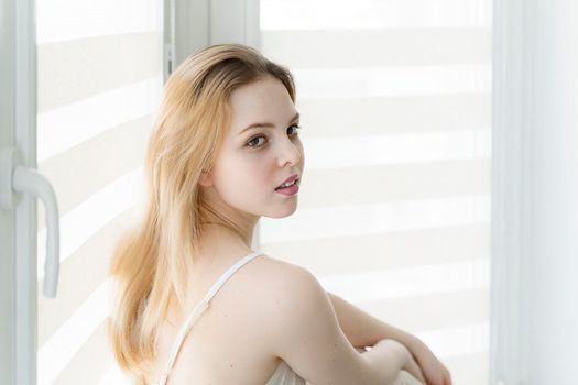 beautiful young woman sits by the window in a white room with her back half-turned.