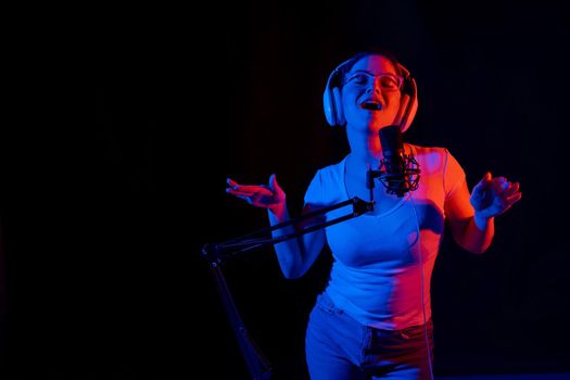 Caucasian woman in glasses and headphones sings into a microphone in neon light on a black background. An emotional girl is recording a song in a recording studio.