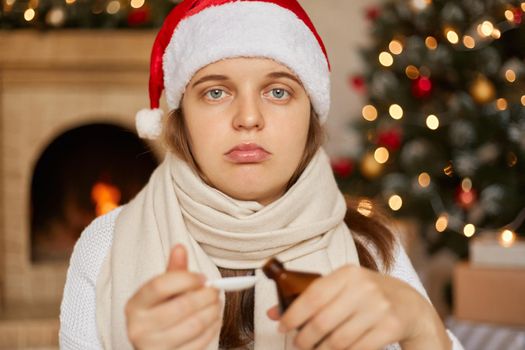 Young Caucasian woman in christmas hat with syrup from coughing, sick due virus or infection, looks at camera with upset expression, wrapped with war scarf, poses in room with New Year decorations