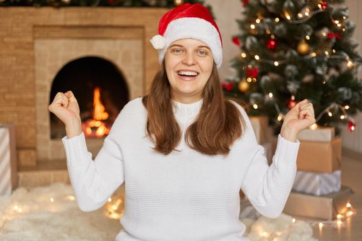 Excited young European Santa girl in warm sweater and Christmas hat, Happy New Year celebration, lady clenching fists like winner, looks at camera with joyful expression.