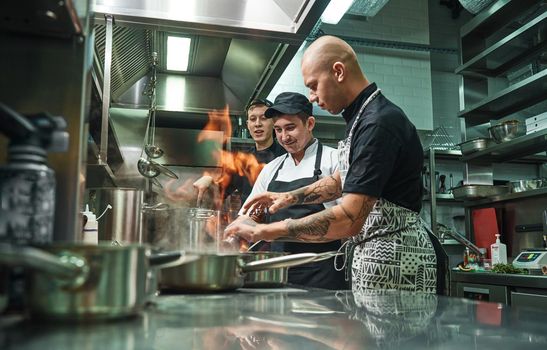 Extreme cooking. Profesional chef teaching his two young trainees how how to flambe food safely. Restaurant kitchen. Cooking process