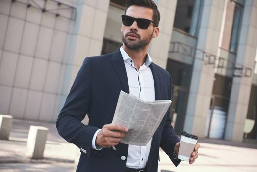 Confident businessman. Happy young businessman reading newspaper outside office building