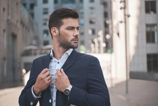 Confident businessman. Confident young man in full suit adjusting his sleeve and looking away while standing outdoors with cityscape in the background