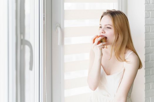 beautiful teen girl in a white dress eats an apple by the window in a bright room