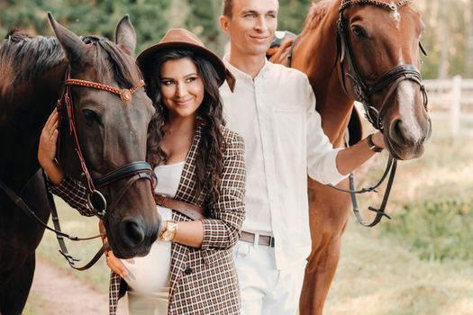 a pregnant girl in a hat and her husband in white clothes stand next to horses in the forest in nature.Stylish pregnant woman with a man with horses.Family