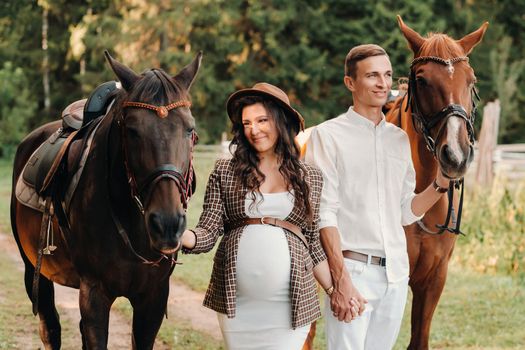 A pregnant woman in a hat with a man in white clothes walking with horses in nature. A family waiting for a child walks in the woods.