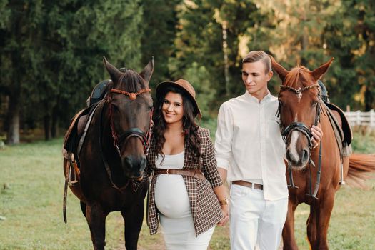 a pregnant girl in a hat and her husband in white clothes stand next to horses in the forest in nature.Stylish pregnant woman with a man with horses.Family