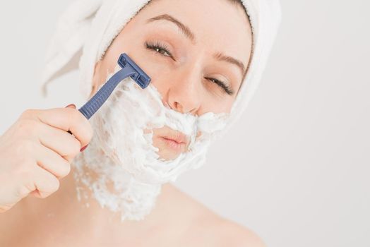 Cheerful caucasian woman with a towel on her head and shaving foam on her face holds a razor on a white background.