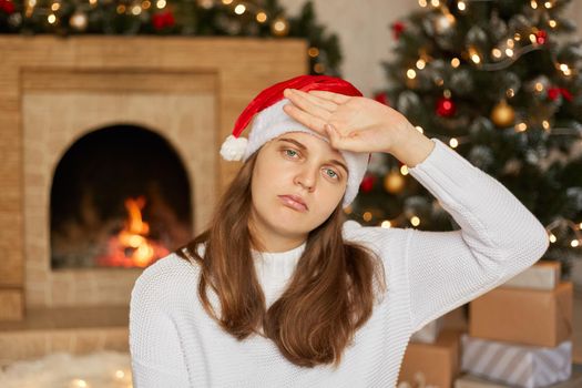 Young beautiful woman wears Santa hat and sweater, touching forehead for illness and fever, flu and cold, virus sick, looks at camera with sad expression, posing with x-mas decoration on background.