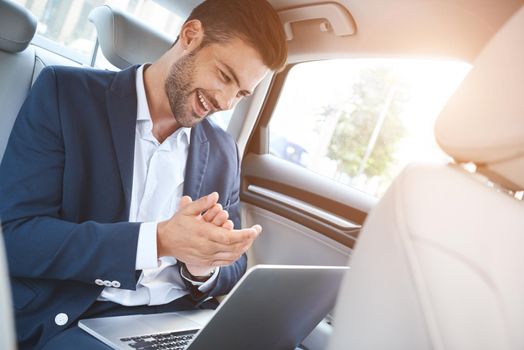 A young man is sitting in the car and enjoying the successful transaction. He looks at the laptop display, which is on his lap