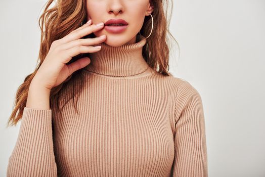 Close up of young beautiful girl with brown hair, wearing beige turtleneck and touching her face, isolated over white background. Full lips, elegant fingers, nice hair.