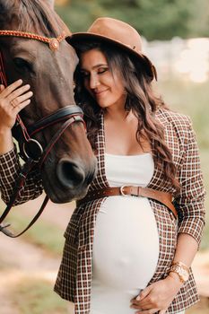 pregnant girl with a big belly in a hat next to horses in the forest in nature.A stylish pregnant woman in a white dress and brown jacket with horses