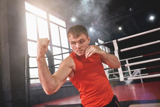 Focused muscular athlete in sports clothing throwing uppercut. Young man boxing with shadow while standing opposite colored boxing ring