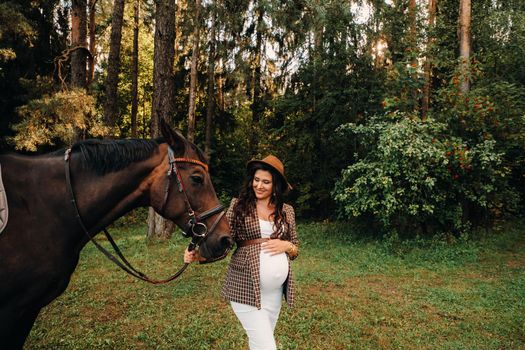 pregnant girl with a big belly in a hat next to horses in the forest in nature.A stylish pregnant woman in a white dress and brown jacket with horses