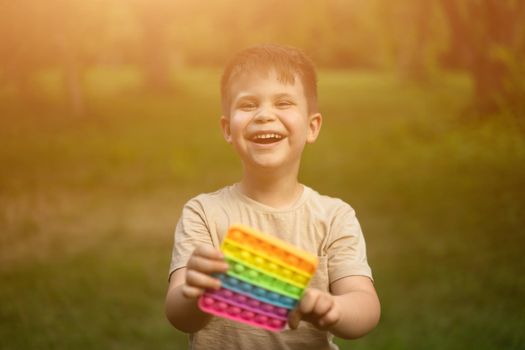 A happy little boy with Pop It enjoys of sun rays in the summer park . A child of 7 years old with a puzzle in his hands. Childhood happiness concept. Blurred background. High quality photo
