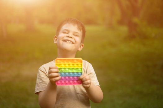 Gigglet Boy Has Fun With His Colorful Pop It and Sunbathes in the Bright Summer Sun. Young One Smiling for a Sun in the Summer Square. Trees and Grass Background. High quality photo