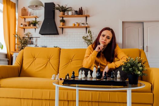 Young upset woman playing chess sitting on sofa. Distressed female plays in logical board game with herself in room