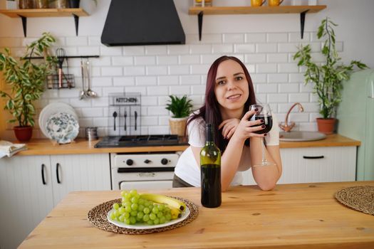 Relaxed young woman poses, standing at kitchen table. Adult brunette resting with alcohol and fruits in kitchen