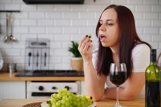 Young woman eats green grape in kitchen. Adult female resting with fruits and alcohol in her weekend
