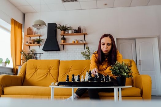 Young woman sitting on yellow sofa and playing chess in room. Female playing in logical board game with herself