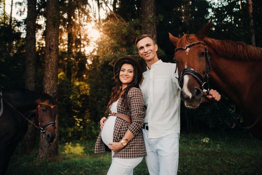 a pregnant girl in a hat and a man in white clothes stand next to horses in the forest in nature.Stylish pregnant woman with her husband with horses.Married couple