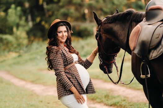 pregnant girl with a big belly in a hat next to horses in the forest in nature.Stylish pregnant woman in the brown dress with the horses