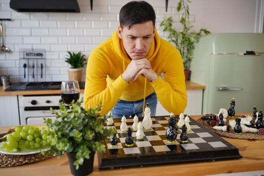 Young man playing chess on kitchen table. Male plays in logical board game with himself, standing in kitchen