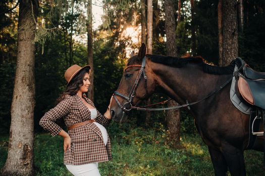 pregnant girl with a big belly in a hat next to horses in the forest in nature.A stylish pregnant woman in a white dress and brown jacket with horses