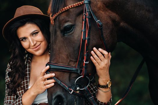 pregnant girl with a big belly in a hat next to horses in the forest in nature.Stylish girl in white clothes and a brown jacket