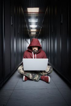 The hacker in a hood sits with the computer among servers, the burglar. Storage of data, cloudy services, system breaking, attack of hackers. High quality photo