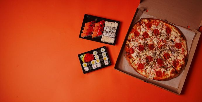 Close up of appetizing sushi in containers and large pizza in box on orange background. Pizza and set of tasty sushi rolls with different ingredients