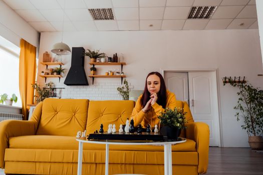 Young woman sitting on yellow sofa and playing chess in room. Female playing in logical board game with herself