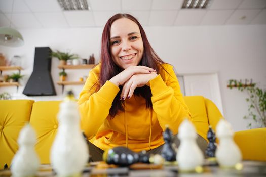 Young woman sitting on yellow sofa and playing chess in room. Female playing in logical board game with herself