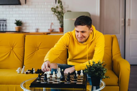 Young man sitting on yellow sofa and playing chess in room. Male playing in logical board game with himself