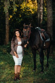 pregnant girl with a big belly in a hat next to horses in the forest in nature.Stylish girl in white clothes and a brown jacket