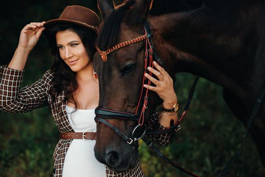 pregnant girl with a big belly in a hat next to horses in the forest in nature.Stylish girl in white clothes and a brown jacket