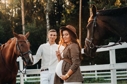 a pregnant girl in a hat and a man in white clothes stand next to horses near a white fence.Stylish pregnant woman with a man with horses.Married couple