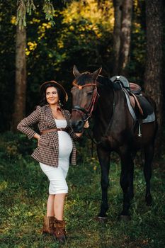 pregnant girl with a big belly in a hat next to horses in the forest in nature.Stylish girl in white clothes and a brown jacket