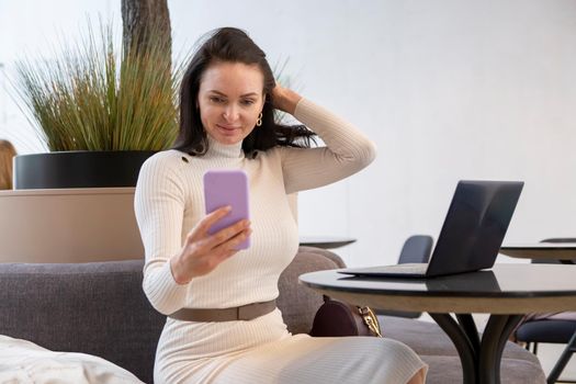 beautiful caucasian woman makes selfie on a smartphone sitting at a table with a laptop. a blogger creates content, a blog author works in a cafe or coworking space.
