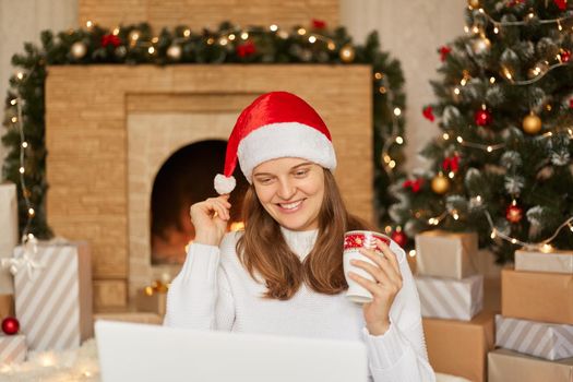 Young woman congratulating with Christmas her close people while sitting on floor in front of tree with her laptop, having happy expression, touching her pom pom from santa hat.