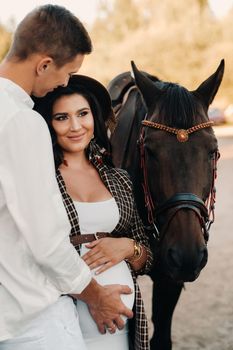 a pregnant girl in a hat and a man in white clothes stand next to horses near a white fence.Stylish pregnant woman with a man with horses.Married couple