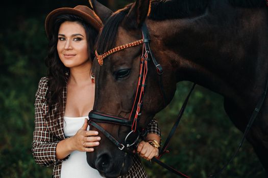 pregnant girl with a big belly in a hat next to horses in the forest in nature.Stylish girl in white clothes and a brown jacket