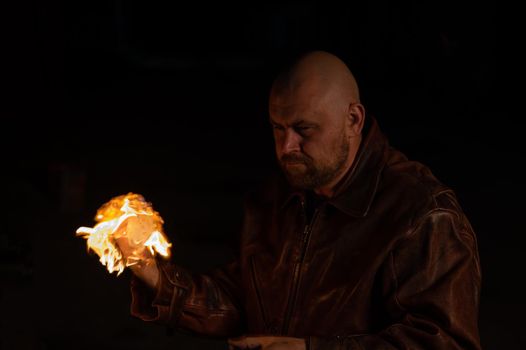 A bald man in a leather jacket holds a burning ball in his hand