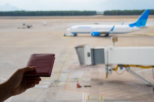 A woman holds a passport against the background of an airplane at the airport. The faceless girl is waiting for her flight and looks at the airfield through the window