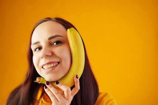 Portrait of young woman with banana on yellow background. Funny female in yellow hoodie posing wth fruit