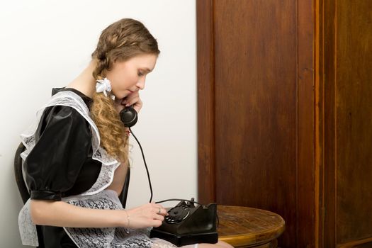 Emotional pretty school girl talking by vintage phone. Side view of girl student with braided hairstyle wearing retro USSR style school uniform posing in room with old fashioned interior