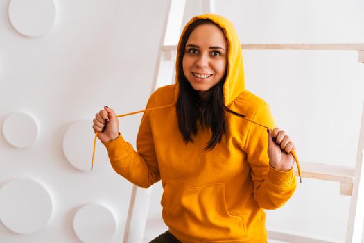 Portrait of young woman posing on white background. Pretty brunette in yellow hoodie near stepladder on background of white wall