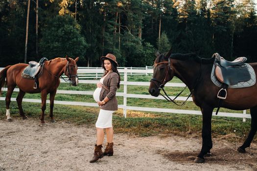 a pregnant girl with a big belly in a hat next to horses near a paddock in nature.Stylish pregnant woman in a brown dress with horses