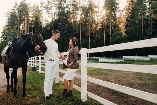 a pregnant girl in a hat and a man in white clothes stand next to horses near a white fence.Stylish pregnant woman with a man with horses.Married couple