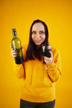 Relaxed young woman poses with bottle and glass of red wine on yellow background. Adult happy brunette holds alcohol in hands